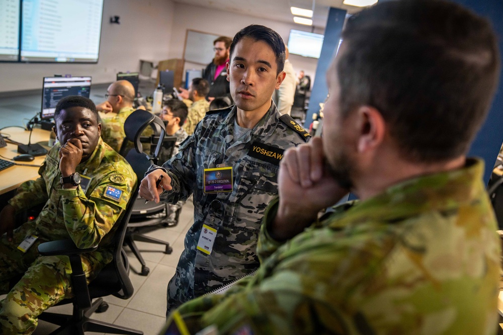 Military personnel from what appears to be multiple nations are gathered in a command or operations center environment, engaged in discussion and briefing. A central figure wearing a name tag reading 'YOSHIZAWA' and a badge labeled 'Translator' is actively speaking and gesturing to colleagues. The setting suggests a joint military exercise or coalition operations planning session.