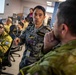Military personnel from what appears to be multiple nations are gathered in a command or operations center environment, engaged in discussion and briefing. A central figure wearing a name tag reading 'YOSHIZAWA' and a badge labeled 'Translator' is actively speaking and gesturing to colleagues. The setting suggests a joint military exercise or coalition operations planning session.