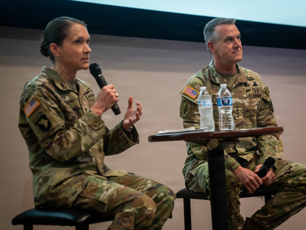 Two U.S. Army officers in OCP (Operational Camouflage Pattern) uniforms are seated on a stage during what appears to be a panel discussion or briefing. The female officer on the left is speaking into a handheld microphone while gesturing with her hands. The male officer on the right, who displays three-star rank insignia, is seated attentively beside a small table with water bottles.