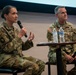 Two U.S. Army officers in OCP (Operational Camouflage Pattern) uniforms are seated on a stage during what appears to be a panel discussion or briefing. The female officer on the left is speaking into a handheld microphone while gesturing with her hands. The male officer on the right, who displays three-star rank insignia, is seated attentively beside a small table with water bottles.