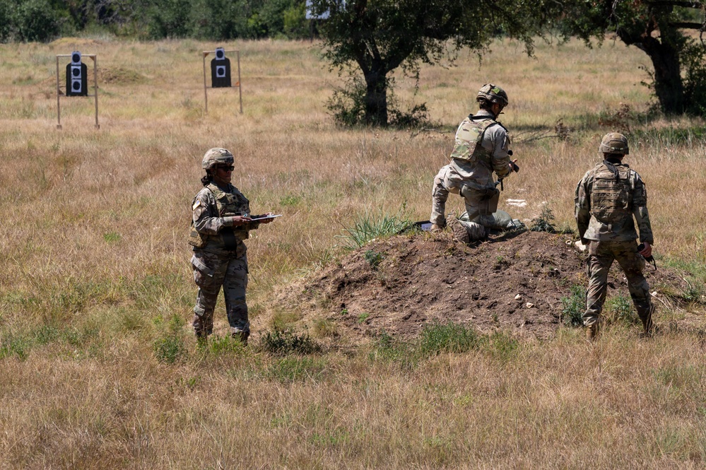 Three military personnel in camouflage uniforms and helmets are on an outdoor shooting range with dry grassland terrain. One soldier kneels on a dirt mound in a firing position, one stands nearby observing, and a third holds a clipboard likely scoring or evaluating performance. Two silhouette targets are visible downrange in the background.