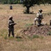 Three military personnel in camouflage uniforms and helmets are on an outdoor shooting range with dry grassland terrain. One soldier kneels on a dirt mound in a firing position, one stands nearby observing, and a third holds a clipboard likely scoring or evaluating performance. Two silhouette targets are visible downrange in the background.