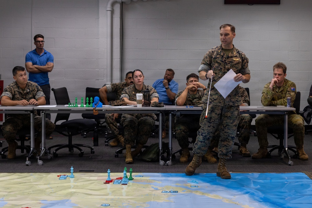 A military briefing or war-game exercise is taking place in an indoor classroom or conference room setting. A standing service member in US Marine Corps camouflage uniform is presenting to a group of seated military personnel using a pointer and papers, with a large floor map displaying colored game pieces. The audience includes personnel in US Marine Corps uniforms and at least one individual in Australian Defence Force camouflage, suggesting a multinational exercise.