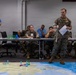 A military briefing or war-game exercise is taking place in an indoor classroom or conference room setting. A standing service member in US Marine Corps camouflage uniform is presenting to a group of seated military personnel using a pointer and papers, with a large floor map displaying colored game pieces. The audience includes personnel in US Marine Corps uniforms and at least one individual in Australian Defence Force camouflage, suggesting a multinational exercise.