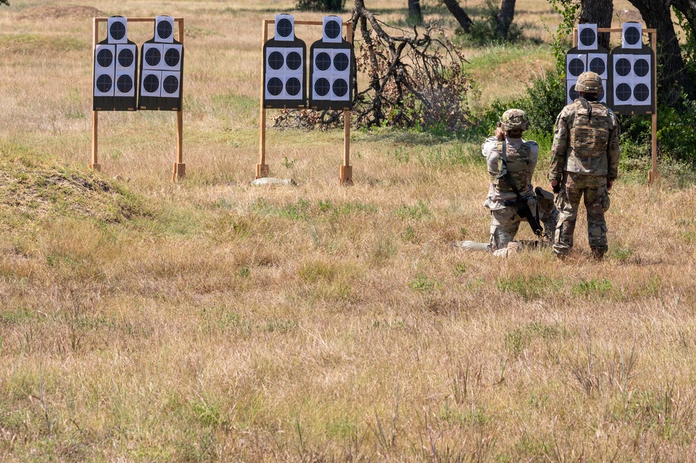 Two military personnel in camouflage uniforms are standing on a shooting range facing multiple paper silhouette targets mounted on wooden frames. One individual appears to be in a kneeling or crouching position while the other stands, suggesting an active training exercise. The setting is an outdoor field range with dry grass and sparse trees in the background.