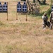 Two military personnel in camouflage uniforms are standing on a shooting range facing multiple paper silhouette targets mounted on wooden frames. One individual appears to be in a kneeling or crouching position while the other stands, suggesting an active training exercise. The setting is an outdoor field range with dry grass and sparse trees in the background.