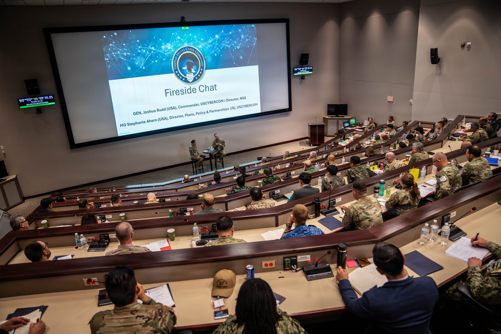 A military briefing or conference is taking place in a tiered auditorium/lecture hall setting. The large projection screen displays a 'Fireside Chat' presentation from USCYBERCOM (United States Cyber Command), featuring GEN Joshua Rudd and MG Stephanie Ahern. The audience consists of numerous military personnel in various uniforms seated in curved rows.