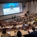 A military briefing or conference is taking place in a tiered auditorium/lecture hall setting. The large projection screen displays a 'Fireside Chat' presentation from USCYBERCOM (United States Cyber Command), featuring GEN Joshua Rudd and MG Stephanie Ahern. The audience consists of numerous military personnel in various uniforms seated in curved rows.