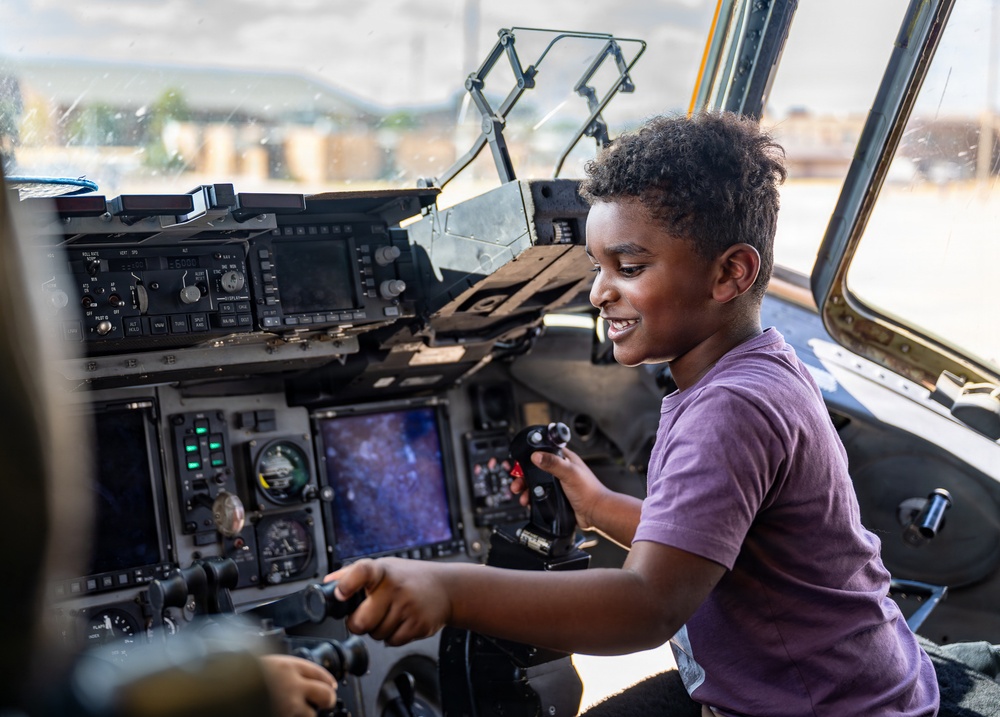 A young boy is seated in the cockpit of what appears to be a military aircraft, smiling as he interacts with the flight controls and instruments. He is holding a joystick/control stick with both hands and appears to be enjoying a guided tour or open-cockpit experience. The aircraft is parked on a tarmac with an outdoor airfield visible through the cockpit windows.