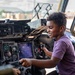 A young boy is seated in the cockpit of what appears to be a military aircraft, smiling as he interacts with the flight controls and instruments. He is holding a joystick/control stick with both hands and appears to be enjoying a guided tour or open-cockpit experience. The aircraft is parked on a tarmac with an outdoor airfield visible through the cockpit windows.