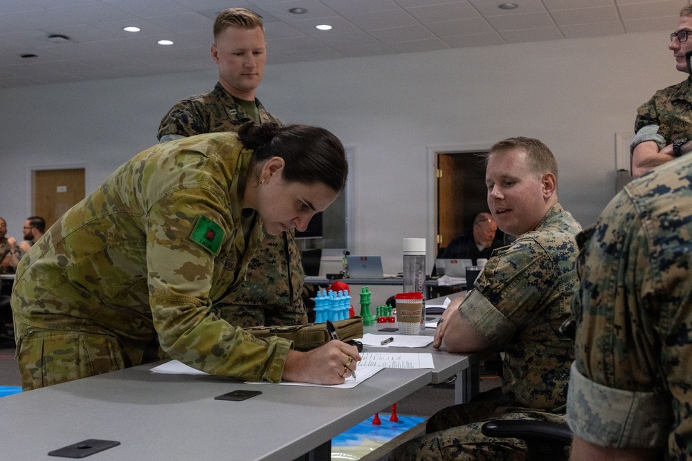 Military personnel from at least two different nations are gathered around a table in what appears to be a training or planning room. A soldier in Australian Army multicam uniform is leaning over a table writing on documents, while U.S. Marines in MARPAT uniforms observe and interact. The table contains colorful game or simulation pieces suggesting a war-gaming or tactical planning exercise.