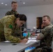 Military personnel from at least two different nations are gathered around a table in what appears to be a training or planning room. A soldier in Australian Army multicam uniform is leaning over a table writing on documents, while U.S. Marines in MARPAT uniforms observe and interact. The table contains colorful game or simulation pieces suggesting a war-gaming or tactical planning exercise.