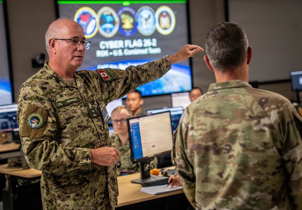 A military briefing or coordination session is taking place in what appears to be a cyber operations center. A Canadian Armed Forces officer (name tag reads 'OSMOND') wearing CADPAT camouflage is gesturing and speaking to a U.S. military personnel in OCP/MultiCam uniform. The background screen displays 'CYBER FLAG 26-2 ROK – U.S. Combined Team,' indicating a joint multinational cyber exercise.