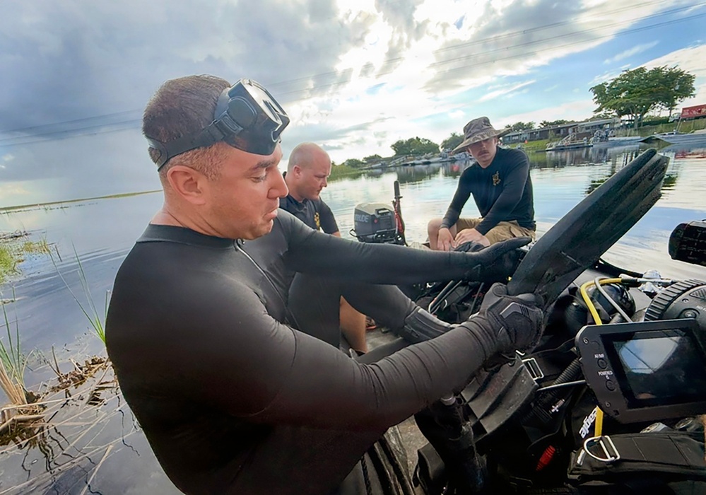 A diver in a black wetsuit with a mask pushed up on his forehead is handling diving equipment near the water's edge. Two other individuals are seated in a small motorboat on a calm waterway, one wearing a camouflage bucket hat and the other in a dark shirt. The setting appears to be a shallow inland waterway or lake, with overcast skies and vegetation visible in the background.