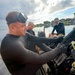 A diver in a black wetsuit with a mask pushed up on his forehead is handling diving equipment near the water's edge. Two other individuals are seated in a small motorboat on a calm waterway, one wearing a camouflage bucket hat and the other in a dark shirt. The setting appears to be a shallow inland waterway or lake, with overcast skies and vegetation visible in the background.