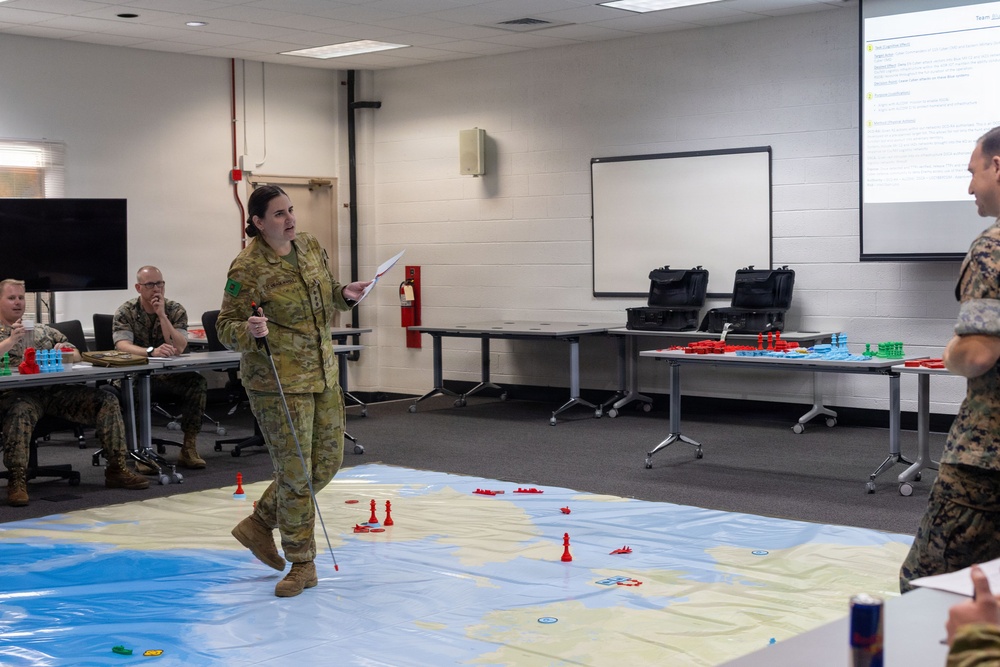 A military briefing or war game exercise is taking place in a classroom or training facility. A soldier in Australian Army multicam uniform is presenting using a large floor map with colored game pieces representing military units or forces. Other military personnel, appearing to wear US Marine Corps uniforms, are observing the briefing.