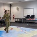 A military briefing or war game exercise is taking place in a classroom or training facility. A soldier in Australian Army multicam uniform is presenting using a large floor map with colored game pieces representing military units or forces. Other military personnel, appearing to wear US Marine Corps uniforms, are observing the briefing.