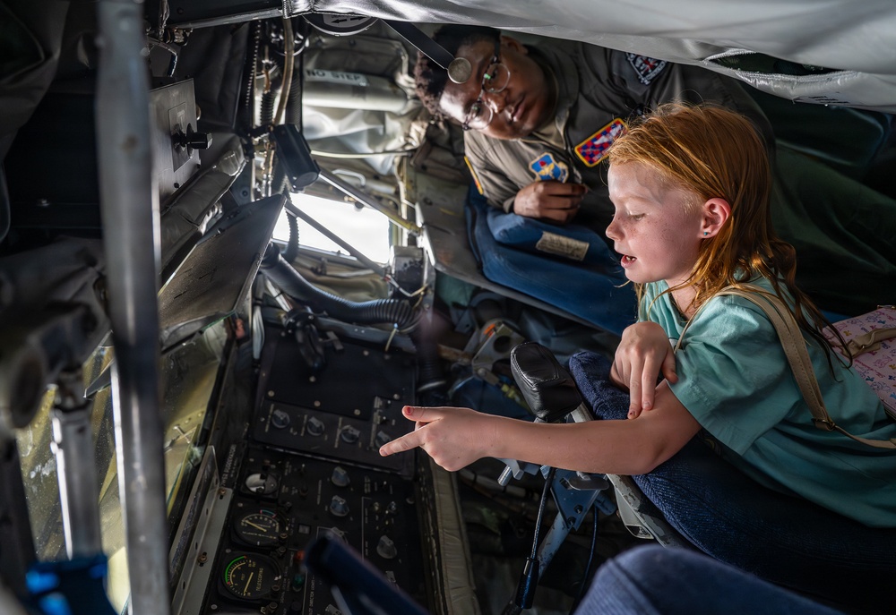 A young red-haired girl is exploring the interior of a military aircraft, likely during an airshow or public tour event. A military service member wearing a uniform with patches is accompanying her and observing as she points at the aircraft's instrumentation panel. The scene captures an educational, hands-on experience inside what appears to be a large military aircraft's boom operator or crew station.