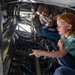 A young red-haired girl is exploring the interior of a military aircraft, likely during an airshow or public tour event. A military service member wearing a uniform with patches is accompanying her and observing as she points at the aircraft's instrumentation panel. The scene captures an educational, hands-on experience inside what appears to be a large military aircraft's boom operator or crew station.