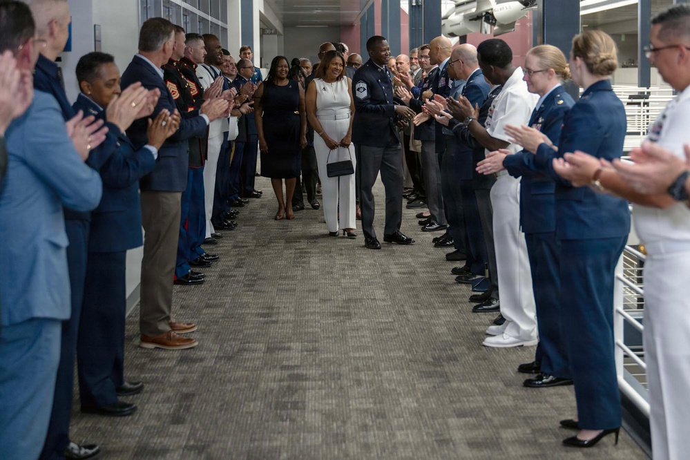 A military honors corridor or 'clapping out' ceremony is taking place in what appears to be a modern indoor hallway. Two civilians in formal attire (one in a black dress, one in white) walk alongside a senior enlisted military member in dress uniform through a gauntlet of applauding service members. The attendees represent multiple military branches, including the Air Force, Marine Corps, and Navy, suggesting a retirement or promotion ceremony.
