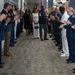 A military honors corridor or 'clapping out' ceremony is taking place in what appears to be a modern indoor hallway. Two civilians in formal attire (one in a black dress, one in white) walk alongside a senior enlisted military member in dress uniform through a gauntlet of applauding service members. The attendees represent multiple military branches, including the Air Force, Marine Corps, and Navy, suggesting a retirement or promotion ceremony.