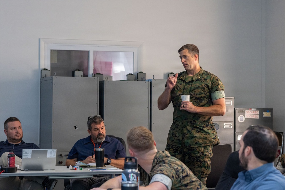 A military briefing or meeting is taking place in an indoor facility. A person in US Marine Corps digital camouflage (MARPAT) uniform is standing and speaking while holding a small paper cup, addressing a group of seated attendees. The audience includes both uniformed military personnel and civilians in casual attire.