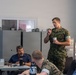A military briefing or meeting is taking place in an indoor facility. A person in US Marine Corps digital camouflage (MARPAT) uniform is standing and speaking while holding a small paper cup, addressing a group of seated attendees. The audience includes both uniformed military personnel and civilians in casual attire.