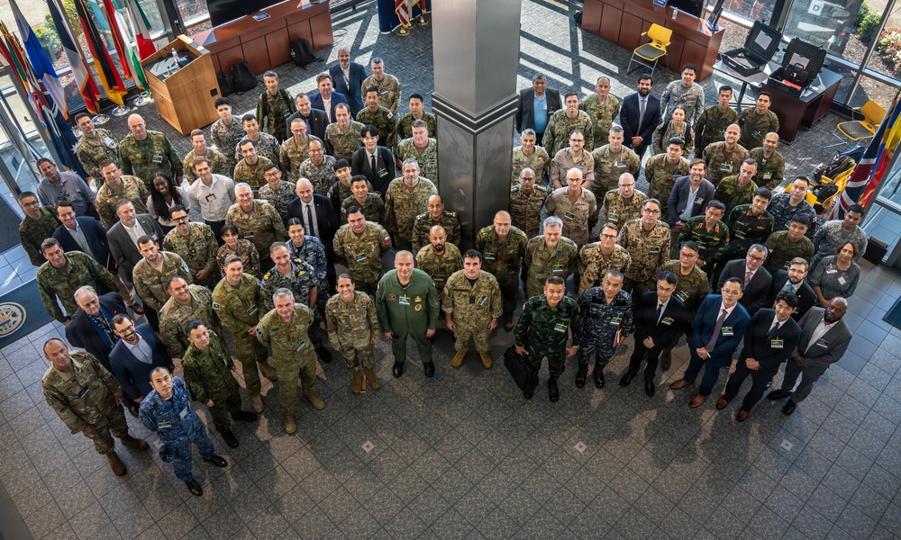 A large group photograph taken from an elevated angle showing approximately 80-100 military personnel and civilians gathered in what appears to be a government or military facility lobby. The group includes individuals wearing various military uniforms from multiple nations as well as civilians in business attire. Multiple national flags are visible in the background, suggesting a multinational or coalition gathering.