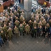 A large group photograph taken from an elevated angle showing approximately 80-100 military personnel and civilians gathered in what appears to be a government or military facility lobby. The group includes individuals wearing various military uniforms from multiple nations as well as civilians in business attire. Multiple national flags are visible in the background, suggesting a multinational or coalition gathering.