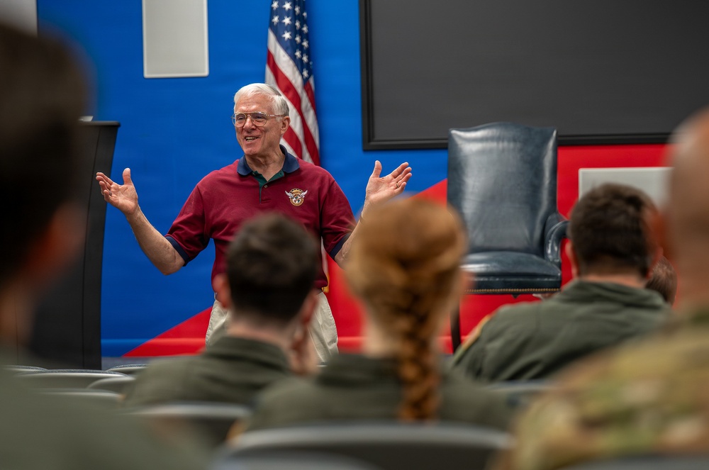 An elderly man wearing a red polo shirt with an emblem is speaking to a seated audience of military personnel in olive/camouflage uniforms. He is gesturing with both hands raised, appearing engaged and animated in his presentation. The setting appears to be a formal briefing or lecture room decorated with blue and red panels and an American flag.