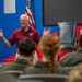 An elderly man wearing a red polo shirt with an emblem is speaking to a seated audience of military personnel in olive/camouflage uniforms. He is gesturing with both hands raised, appearing engaged and animated in his presentation. The setting appears to be a formal briefing or lecture room decorated with blue and red panels and an American flag.