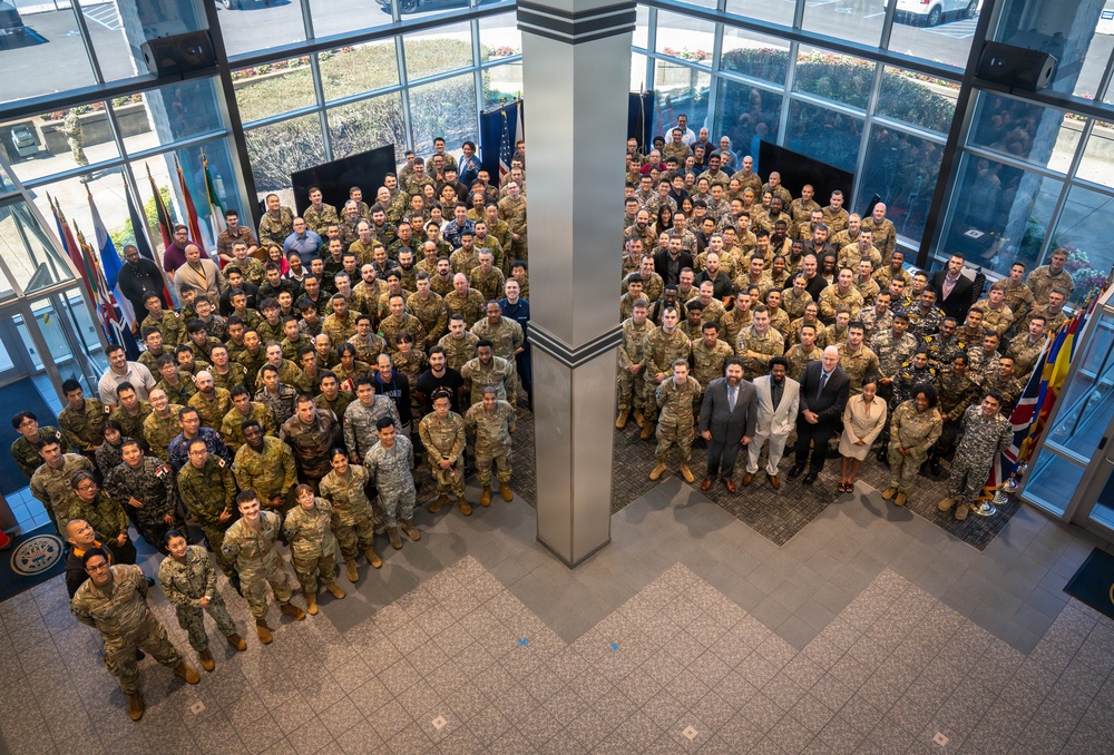 A large group photograph taken from an elevated angle inside a modern building lobby with floor-to-ceiling windows. The group consists predominantly of military personnel in various uniforms, along with a small number of civilians in business attire. Multiple national flags are visible on the left and right sides, suggesting an international or multinational military gathering.