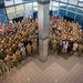 A large group photograph taken from an elevated angle inside a modern building lobby with floor-to-ceiling windows. The group consists predominantly of military personnel in various uniforms, along with a small number of civilians in business attire. Multiple national flags are visible on the left and right sides, suggesting an international or multinational military gathering.