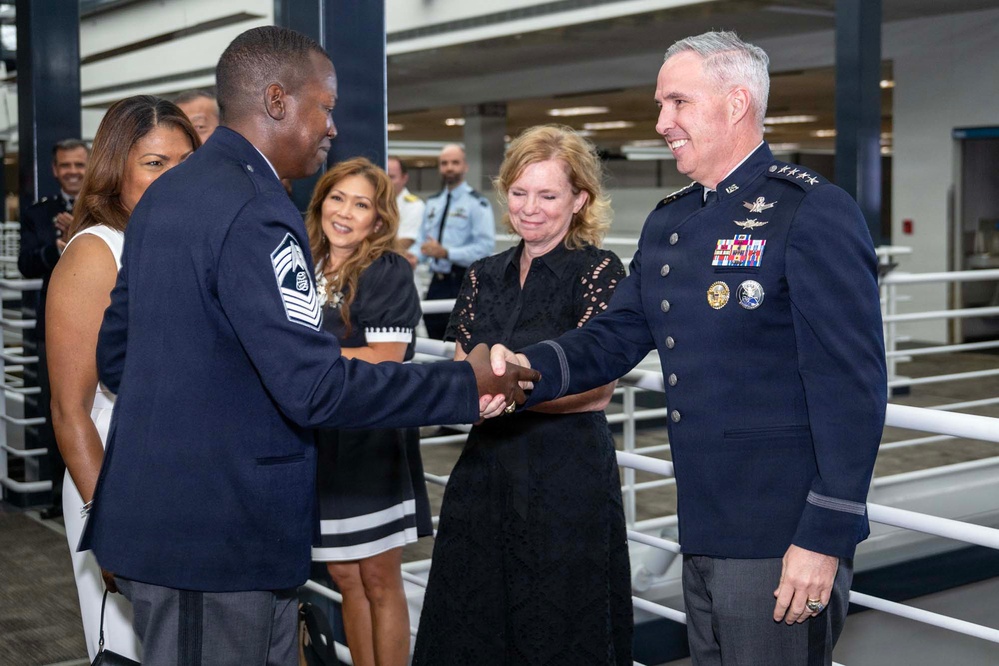 A formal military ceremony is taking place in which a senior enlisted member (Chief Master Sergeant, identifiable by chevron insignia) is shaking hands with a high-ranking officer (General, identifiable by four-star insignia and pilot wings). A woman in a black dress participates in the handshake, suggesting a coin or item presentation. Several onlookers in civilian and military attire observe the exchange in the background.