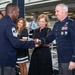 A formal military ceremony is taking place in which a senior enlisted member (Chief Master Sergeant, identifiable by chevron insignia) is shaking hands with a high-ranking officer (General, identifiable by four-star insignia and pilot wings). A woman in a black dress participates in the handshake, suggesting a coin or item presentation. Several onlookers in civilian and military attire observe the exchange in the background.