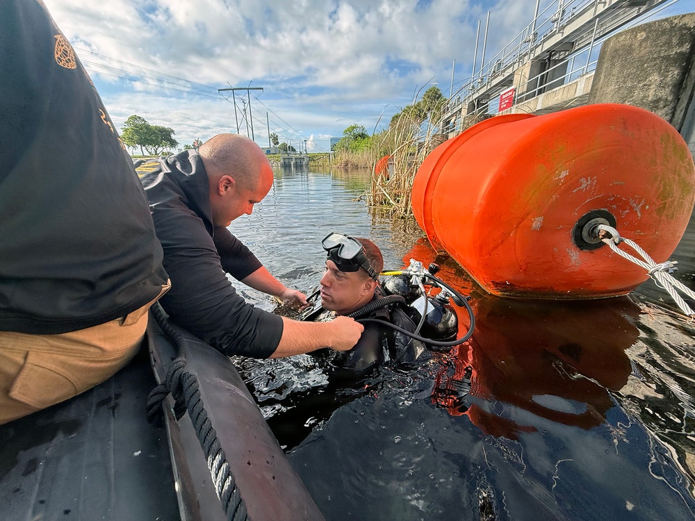 A scuba diver in full black wetsuit and diving gear is in dark water near a large orange buoy, being assisted by a person leaning from what appears to be a boat or dock edge. The setting appears to be a waterway near an industrial or flood control structure. The scene suggests a professional or emergency diving operation.
