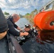 A scuba diver in full black wetsuit and diving gear is in dark water near a large orange buoy, being assisted by a person leaning from what appears to be a boat or dock edge. The setting appears to be a waterway near an industrial or flood control structure. The scene suggests a professional or emergency diving operation.