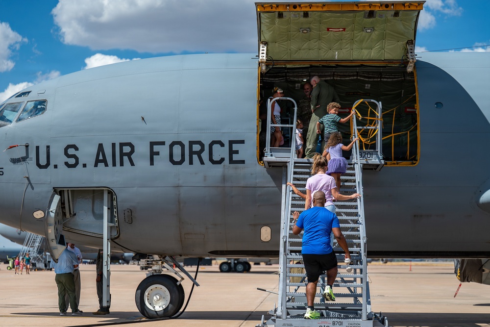 Civilians are boarding a U.S. Air Force aircraft via a portable staircase, likely during an air show or open house event. The large military aircraft displays 'U.S. AIR FORCE' markings on its fuselage, and a side cargo/passenger door is open to allow public access. Multiple adults and children are visible climbing the stairs and entering the aircraft.