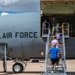 Civilians are boarding a U.S. Air Force aircraft via a portable staircase, likely during an air show or open house event. The large military aircraft displays 'U.S. AIR FORCE' markings on its fuselage, and a side cargo/passenger door is open to allow public access. Multiple adults and children are visible climbing the stairs and entering the aircraft.
