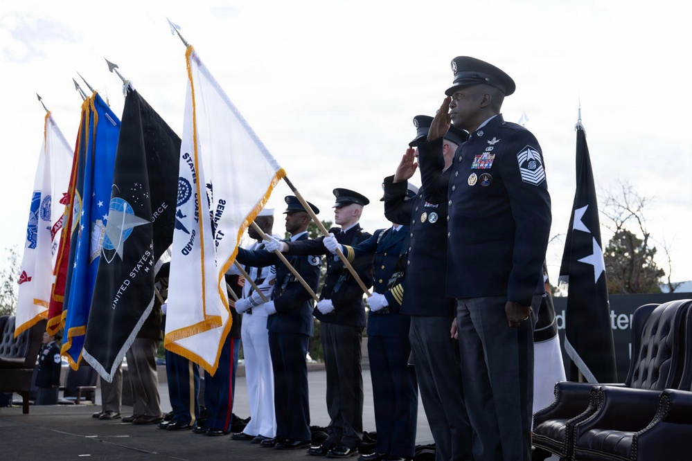 A formal military joint service color guard ceremony is taking place outdoors, with personnel from multiple U.S. military branches presenting their respective service flags. A senior non-commissioned officer in Air Force dress uniform, bearing multiple decorations and a Chief Master Sergeant rank insignia, is prominently saluting in the foreground. The ceremony appears to be an official military event with all branches represented.