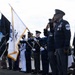 A formal military joint service color guard ceremony is taking place outdoors, with personnel from multiple U.S. military branches presenting their respective service flags. A senior non-commissioned officer in Air Force dress uniform, bearing multiple decorations and a Chief Master Sergeant rank insignia, is prominently saluting in the foreground. The ceremony appears to be an official military event with all branches represented.