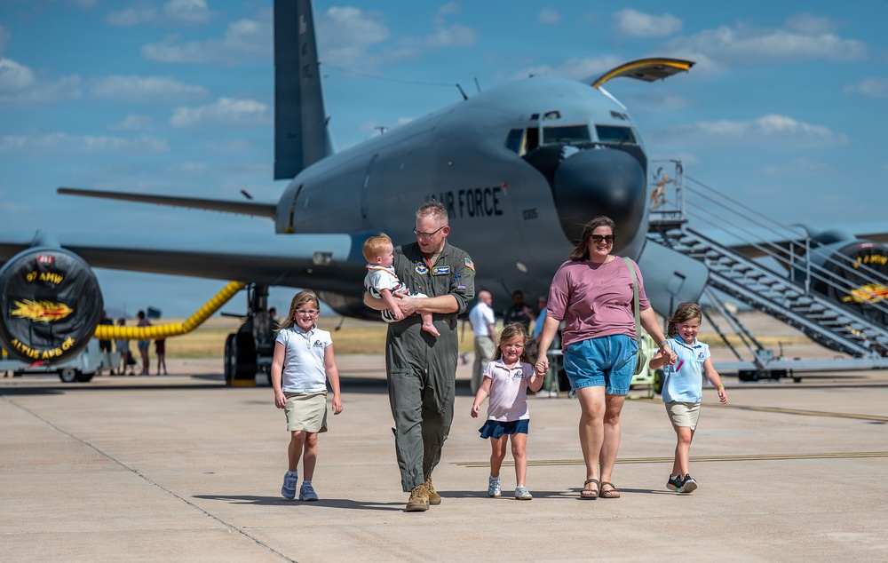 A U.S. Air Force pilot in olive flight suit walks with his family on a flight line tarmac in front of a large military aircraft. The group includes what appears to be a woman and four young children, suggesting a homecoming or family day event at a military air base. The aircraft in the background is identifiable as a KC-135 Stratotanker belonging to the 97th Air Mobility Wing at Altus AFB.