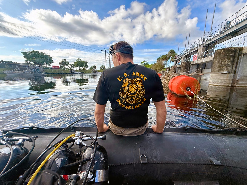 A U.S. Army Deep Sea Diver sits on the edge of an inflatable dive boat near a water control structure or lock, facing away from the camera. The diver is equipped with scuba gear visible in the foreground, and an orange buoy is visible near a concrete dam or floodgate structure to the right. The setting appears to be a Florida waterway, suggested by the palm trees, flat terrain, and tropical vegetation.