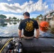 A U.S. Army Deep Sea Diver sits on the edge of an inflatable dive boat near a water control structure or lock, facing away from the camera. The diver is equipped with scuba gear visible in the foreground, and an orange buoy is visible near a concrete dam or floodgate structure to the right. The setting appears to be a Florida waterway, suggested by the palm trees, flat terrain, and tropical vegetation.