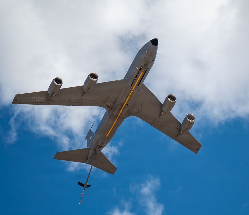 A large military jet aircraft is photographed from directly below, showing its underside as it flies overhead against a partly cloudy blue sky. The aircraft appears to be a US Air Force tanker or transport, identifiable by its four underwing engines, gray paint scheme, and a yellow-striped refueling boom extended from the rear fuselage. The low-angle upward perspective provides a clear view of the aircraft's belly, wings, and tail assembly.