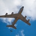 A large military jet aircraft is photographed from directly below, showing its underside as it flies overhead against a partly cloudy blue sky. The aircraft appears to be a US Air Force tanker or transport, identifiable by its four underwing engines, gray paint scheme, and a yellow-striped refueling boom extended from the rear fuselage. The low-angle upward perspective provides a clear view of the aircraft's belly, wings, and tail assembly.