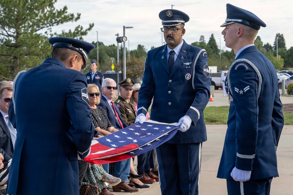 A U.S. Air Force Honor Guard is performing a formal flag-folding ceremony, likely at a military memorial or funeral service. Three uniformed airmen in dress blues with white gloves are carefully folding an American flag while an audience of civilians and military personnel observes. The setting appears to be an outdoor military installation.