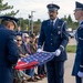 A U.S. Air Force Honor Guard is performing a formal flag-folding ceremony, likely at a military memorial or funeral service. Three uniformed airmen in dress blues with white gloves are carefully folding an American flag while an audience of civilians and military personnel observes. The setting appears to be an outdoor military installation.