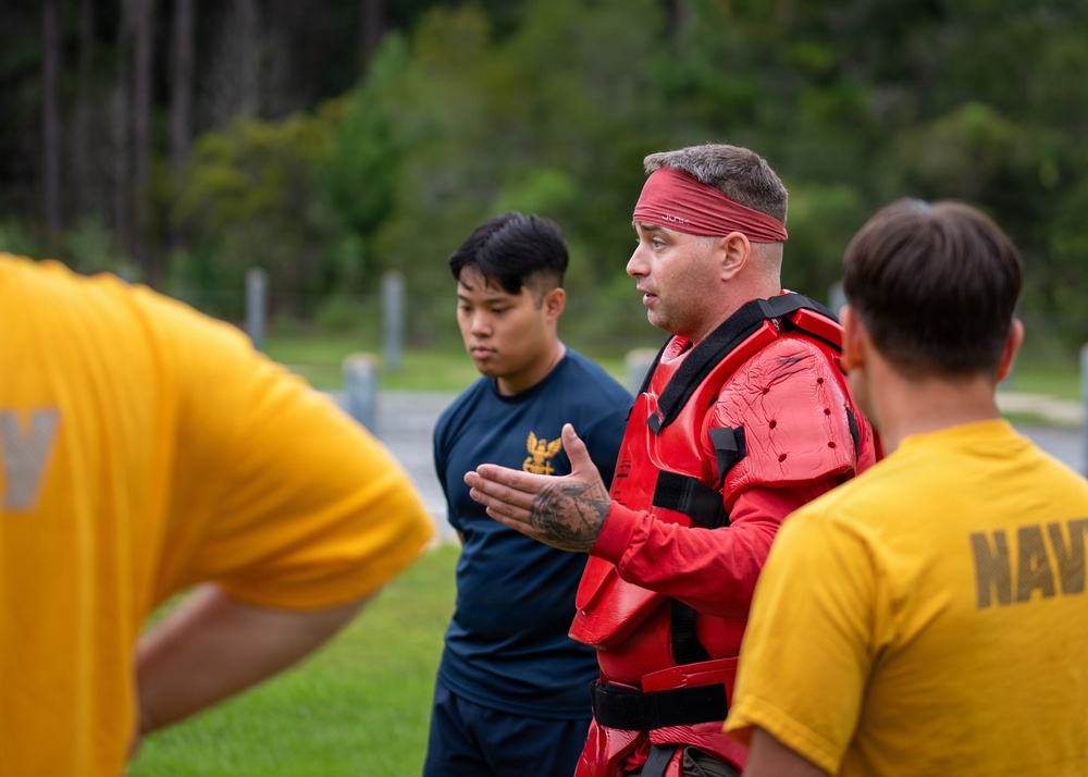 A group of U.S. Navy personnel are gathered outdoors in what appears to be a combatives or self-defense training session. A central figure wearing red protective padding and a red headband appears to be an instructor demonstrating or explaining techniques to trainees in yellow 'NAVY' t-shirts. The setting is an outdoor training area with a wooded background.