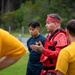 A group of U.S. Navy personnel are gathered outdoors in what appears to be a combatives or self-defense training session. A central figure wearing red protective padding and a red headband appears to be an instructor demonstrating or explaining techniques to trainees in yellow 'NAVY' t-shirts. The setting is an outdoor training area with a wooded background.